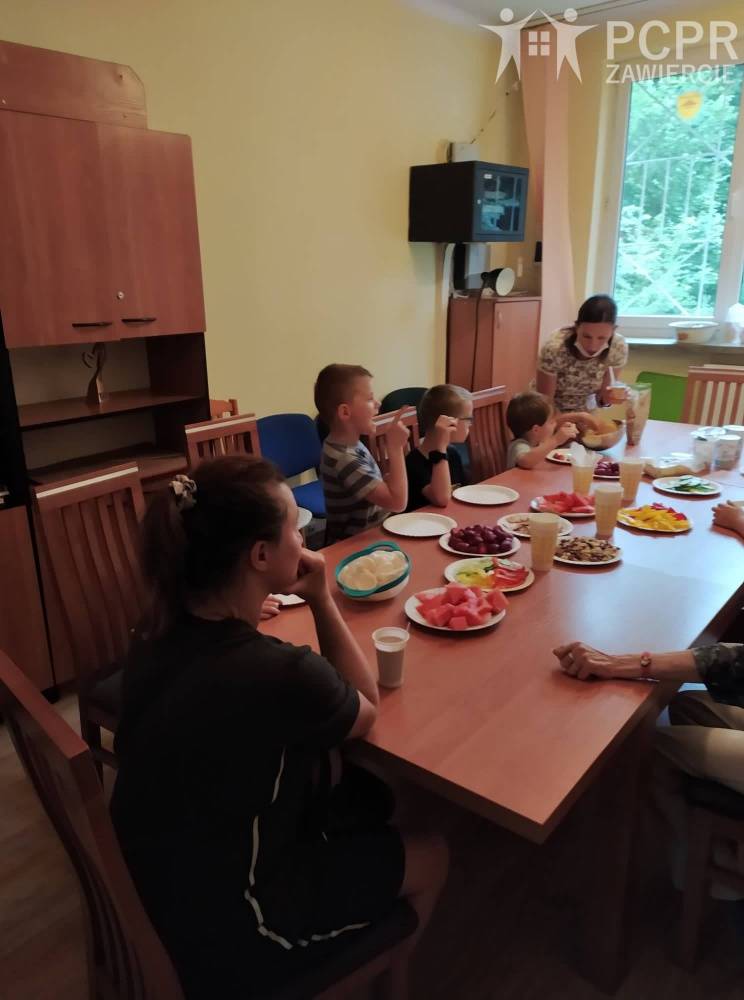 Zdjęcie: Grupa dzieci siedzi przy stole, na którym w pojemnikach poustawiane są pokrojone produkty spożywcze, a kobieta tłumaczy
